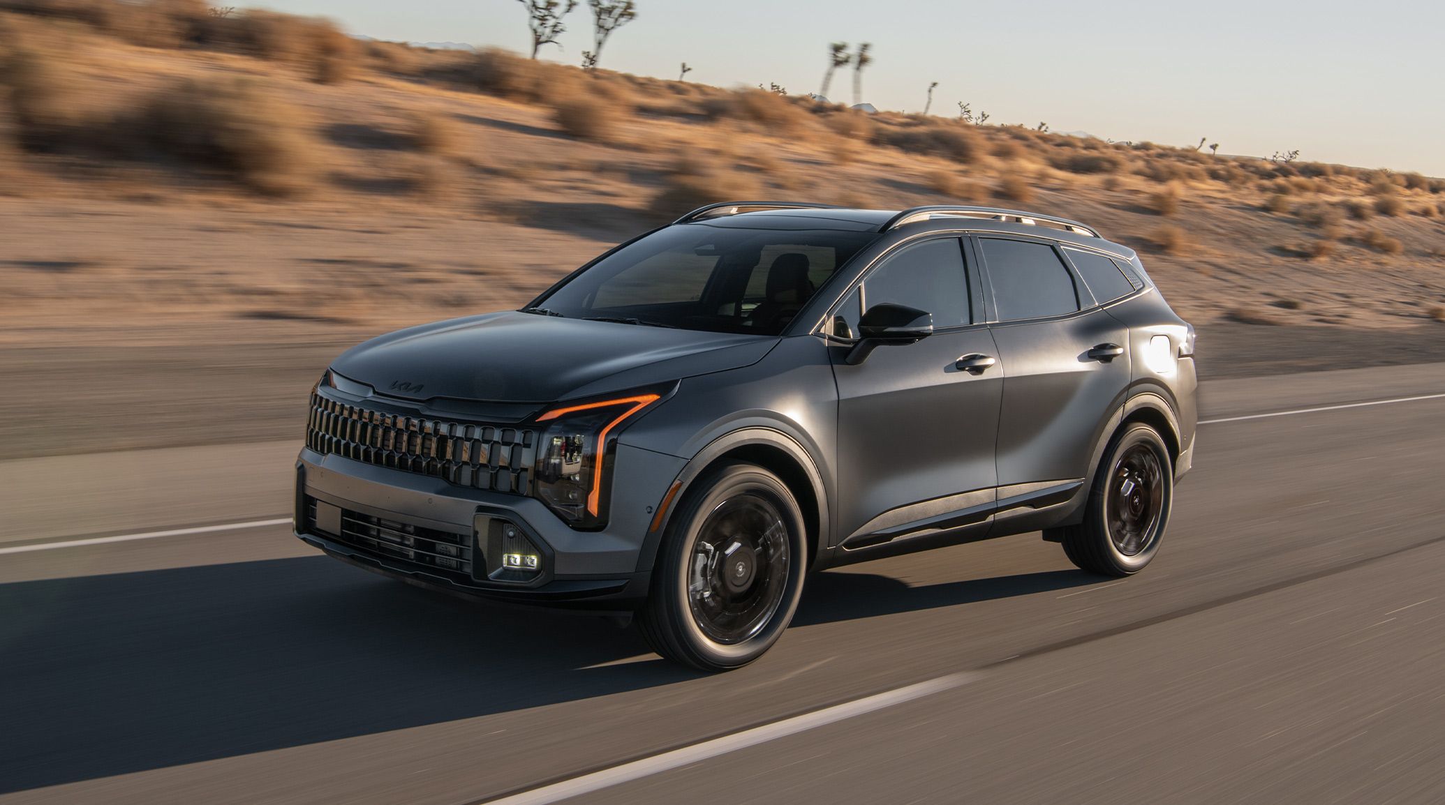 A gray 2027 Kia Sportage Plug-In Hybrid compact SUV is driving along a desert highway, highlighting the driver’s side and sleek design, plus LED amber daytime lights.