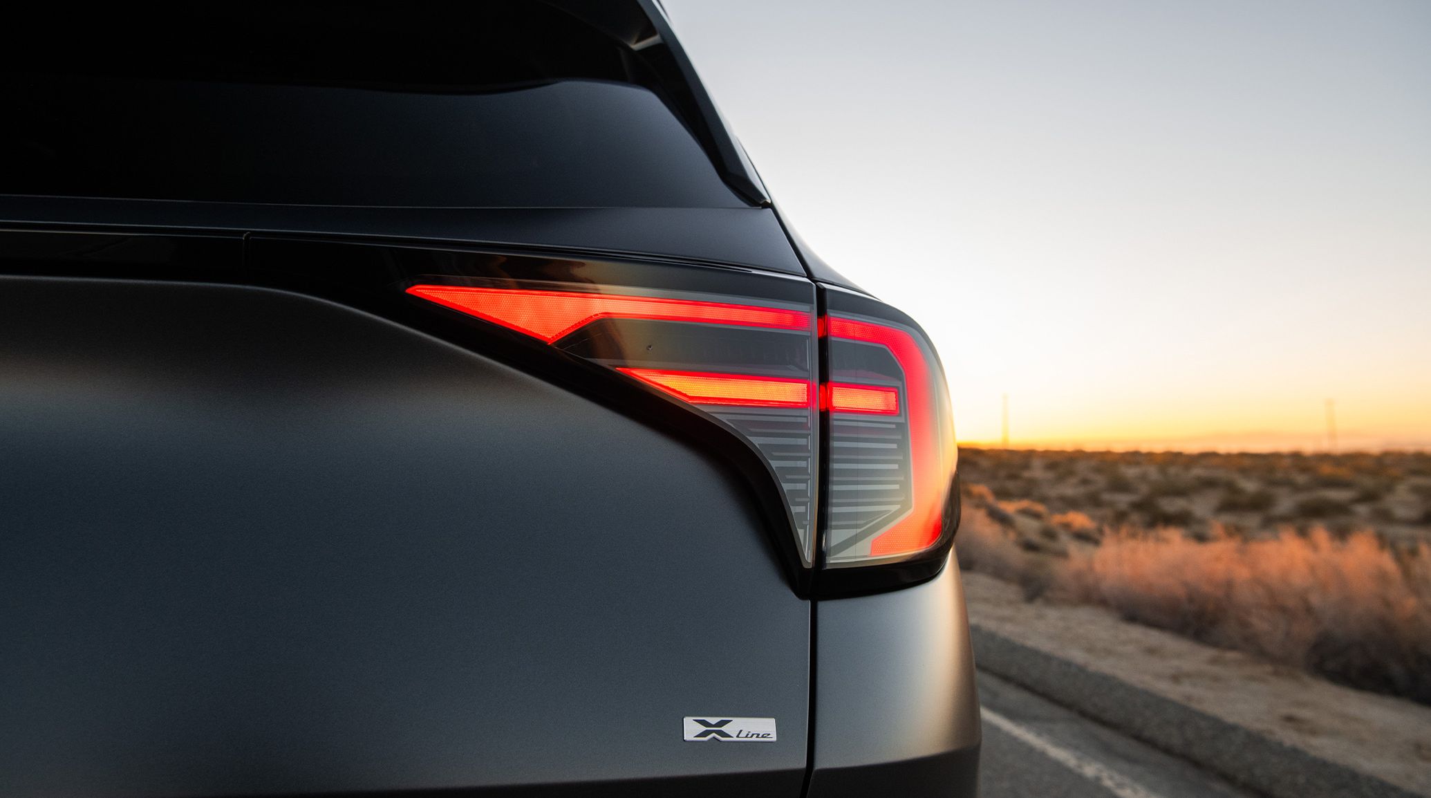 A still shot of the 2027 Kia Sportage Plug-In Hybrid compact SUV in matte gray parked with the trunk closed, showcasing the SUV's sleek LED taillights against a desert backdrop during sunset.