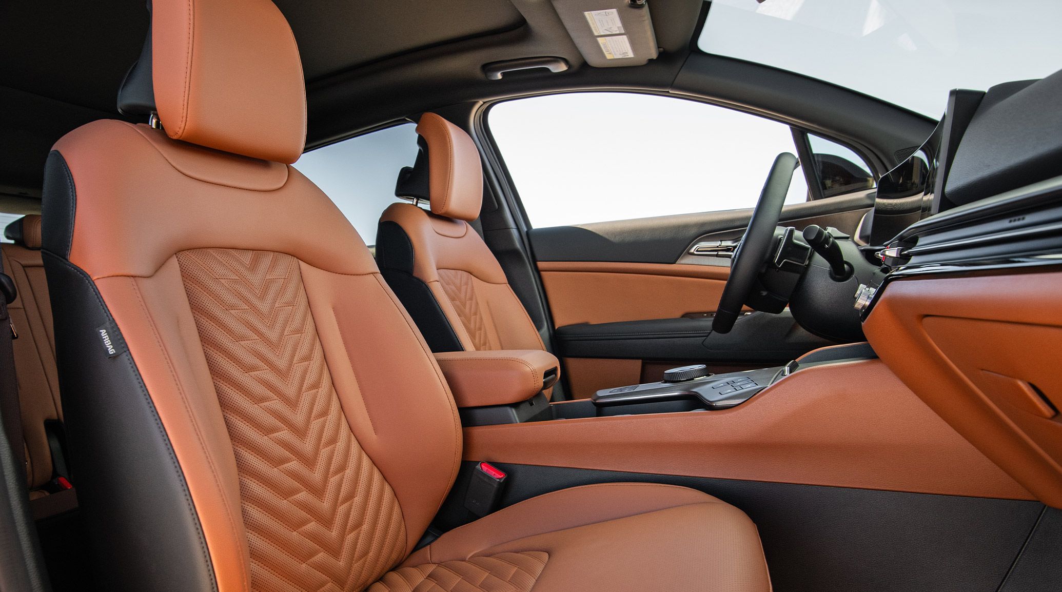 Interior passenger-side view of the 2027 Kia Sportage Plug-In Hybrid in brown and black, highlighting the comfortable front row seats, center console, steering wheel, and modern design. Blue skies are visible through the window.