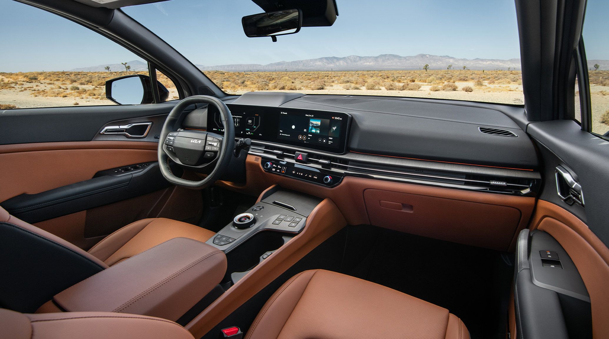 Interior view of the 2027 Kia Sportage Plug-In Hybrid in brown featuring the driver-side steering wheel, digital display with maps and music showing, and the center console with a scenic desert landscape and mountains visible through the windshield.