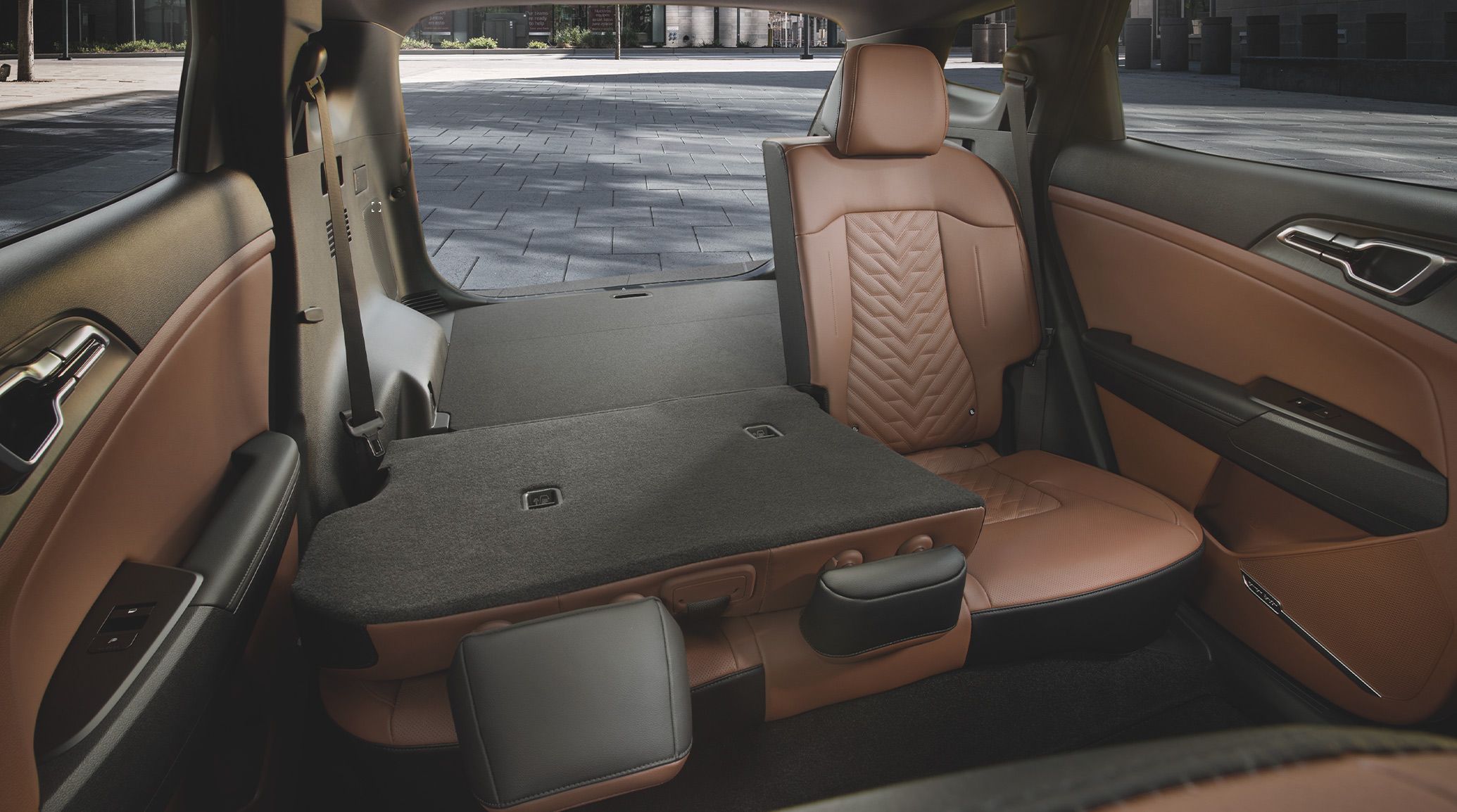 Rear seating and cargo area of the 2027 Kia Sportage Plug‑In Hybrid with 2 seats folded down, showing brown leather upholstery and the spacious cargo area with the trunk open and a city landscape in the background.