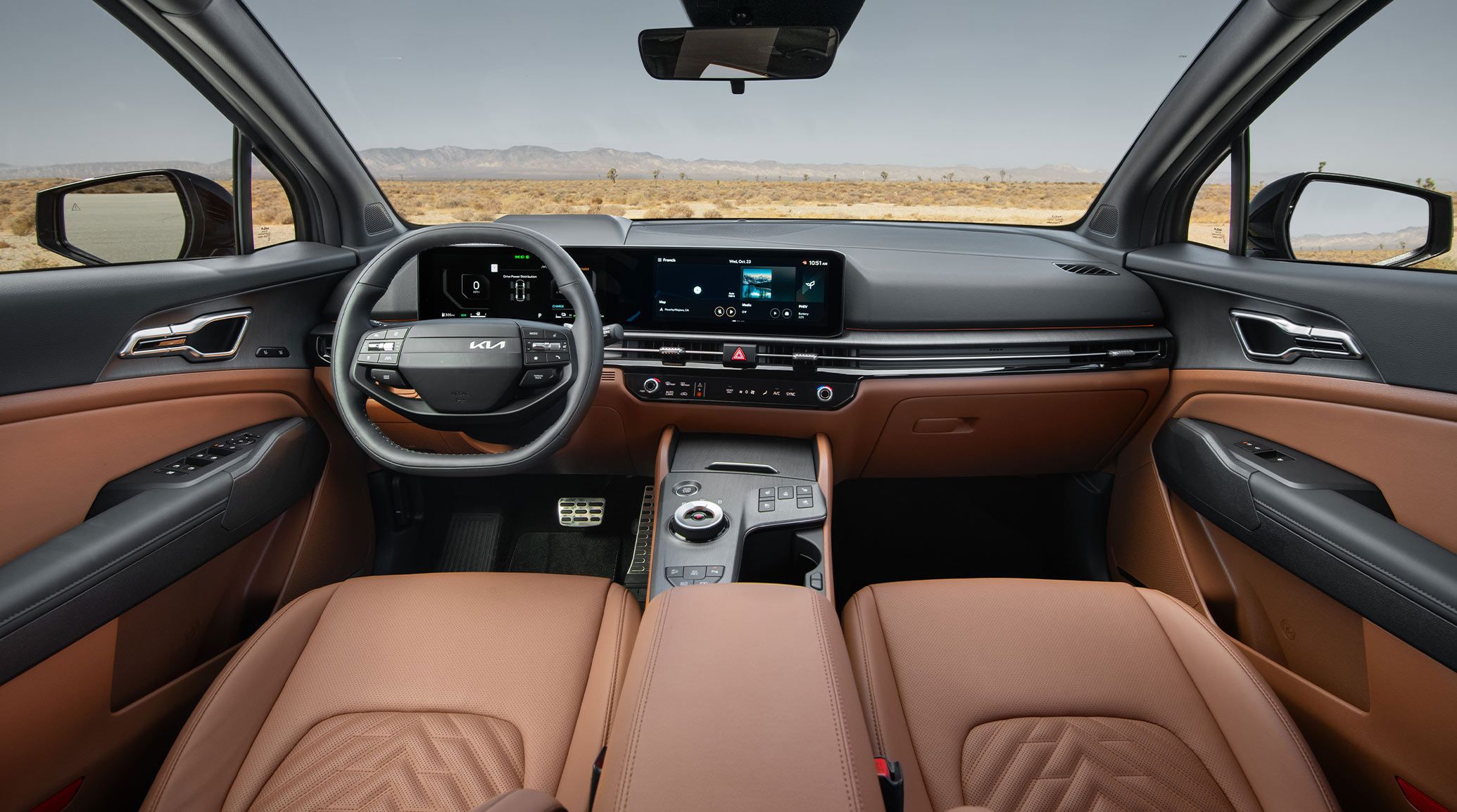 The interior of a Kia vehicle showcasing the dashboard, steering wheel, center console, and other convenient features. Outside, a landscape featuring desert scrub and clouds is visible stretching into the distance.