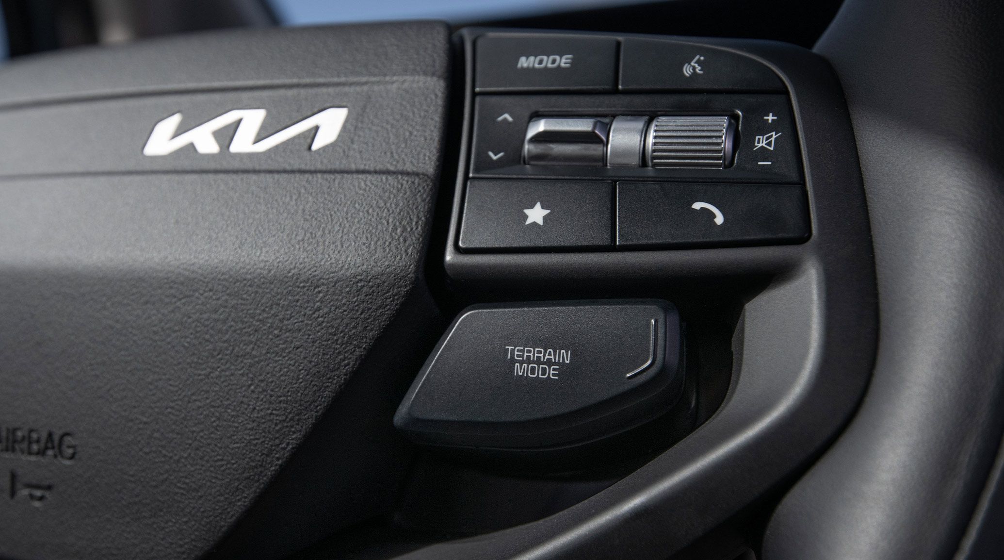 Close-up image of the steering wheel button panel inside the 2027 Kia Sportage SUV, including the Kia logo, mode, voice, star, and phone buttons, an up/down dial and volume control wheel, and a terrain mode button underneath.