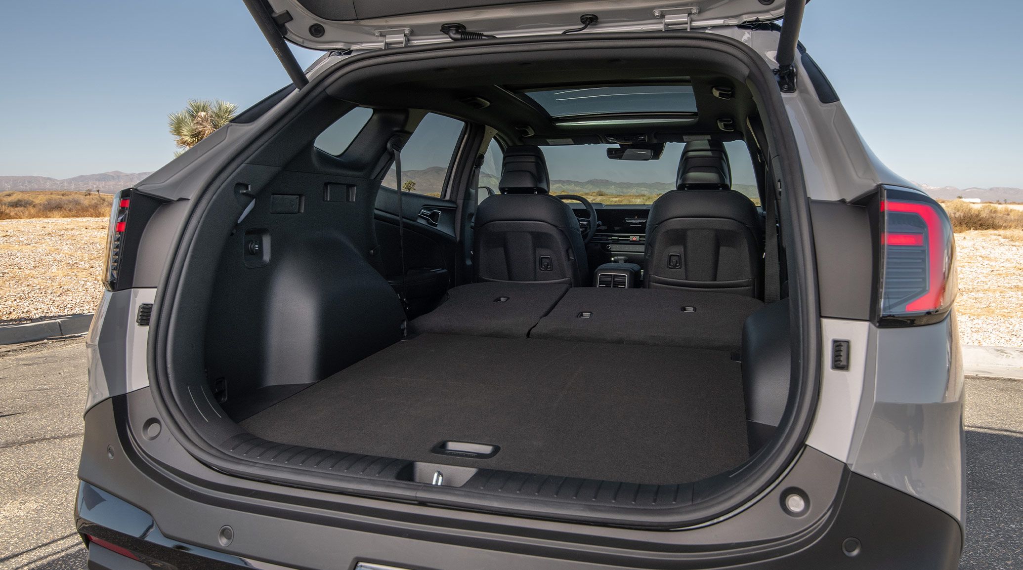 Rear angled photograph of the 2027 Kia Sportage SUV with the trunk door opened up, showing a view of the empty cargo area with the 2nd-row seats folded down. A desert background is visible behind the vehicle.