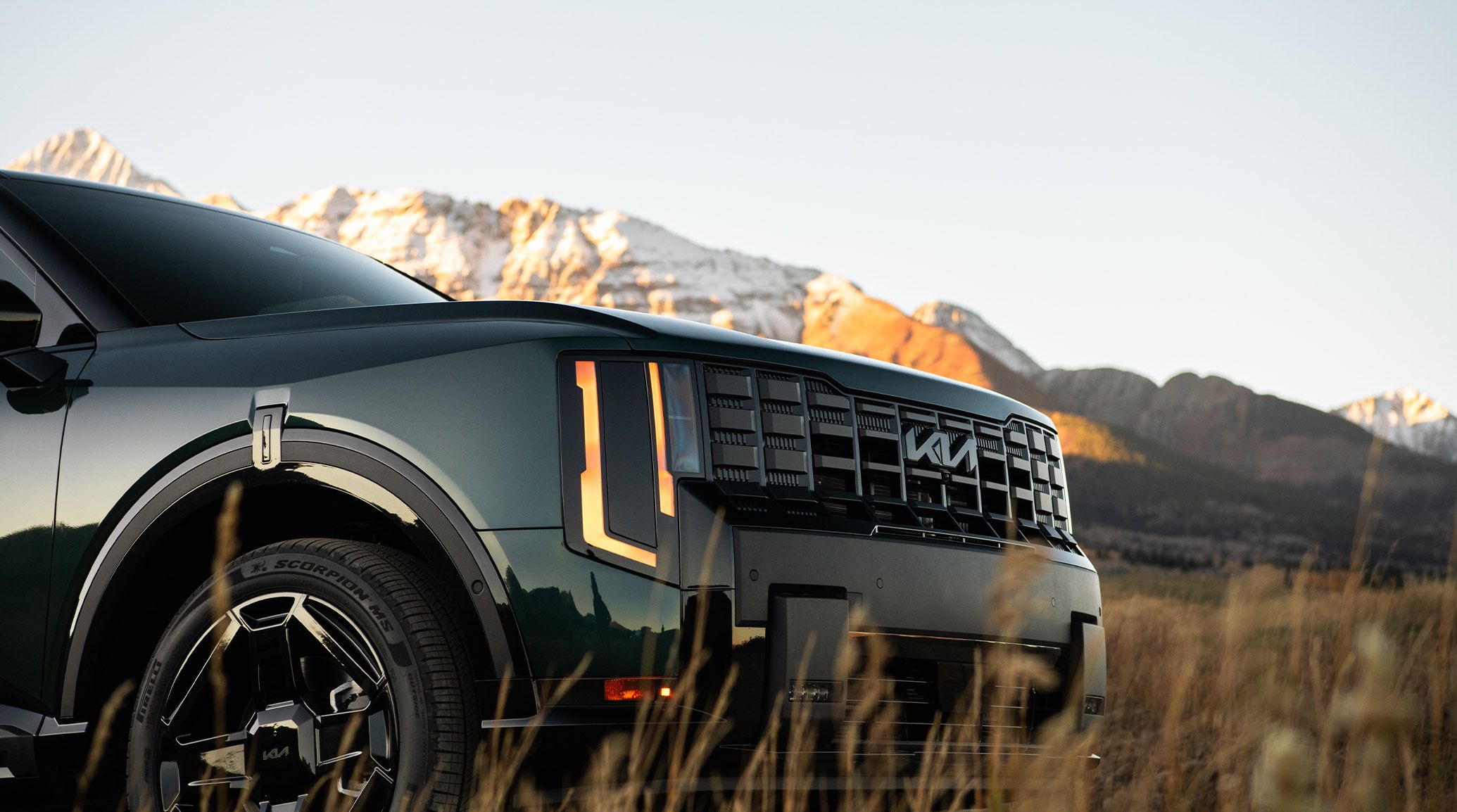 A close-up shot of the 2027 Kia Telluride Hybrid in jade green, highlighting the running lights, bold grille, and all-terrain tires. The SUV is parked on a grassy meadow with snow-capped mountains behind.