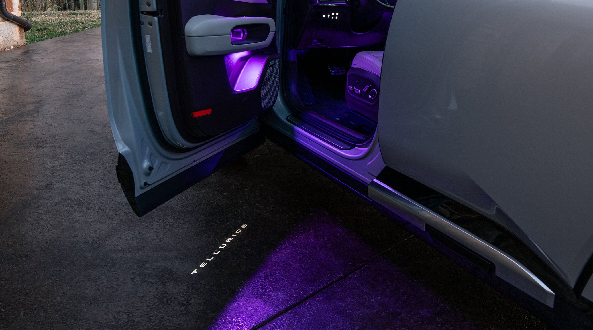 The 2027 Kia Telluride Hybrid in silver with a close-up of the driver-side door open, showcasing the illuminated Telluride logo projected onto the ground. Purple LED interior lighting is visible, highlighting the premium design details.