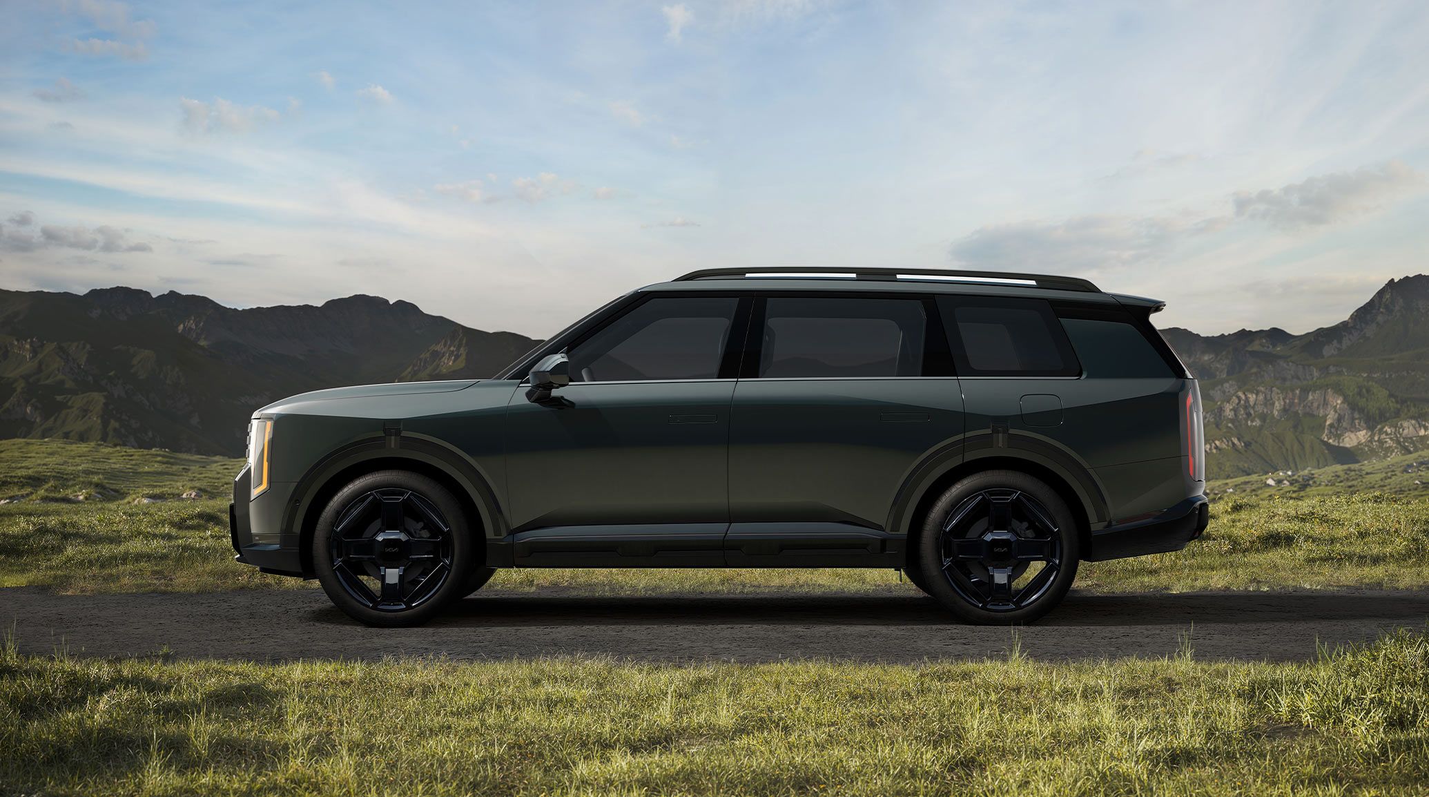 A driver-side view of the 2027 Kia Telluride Hybrid in jade green, parked on a dirt patch with grass and rocky hills around the vehicle, showcasing the SUV’s high utility roof rails and bold exterior design.