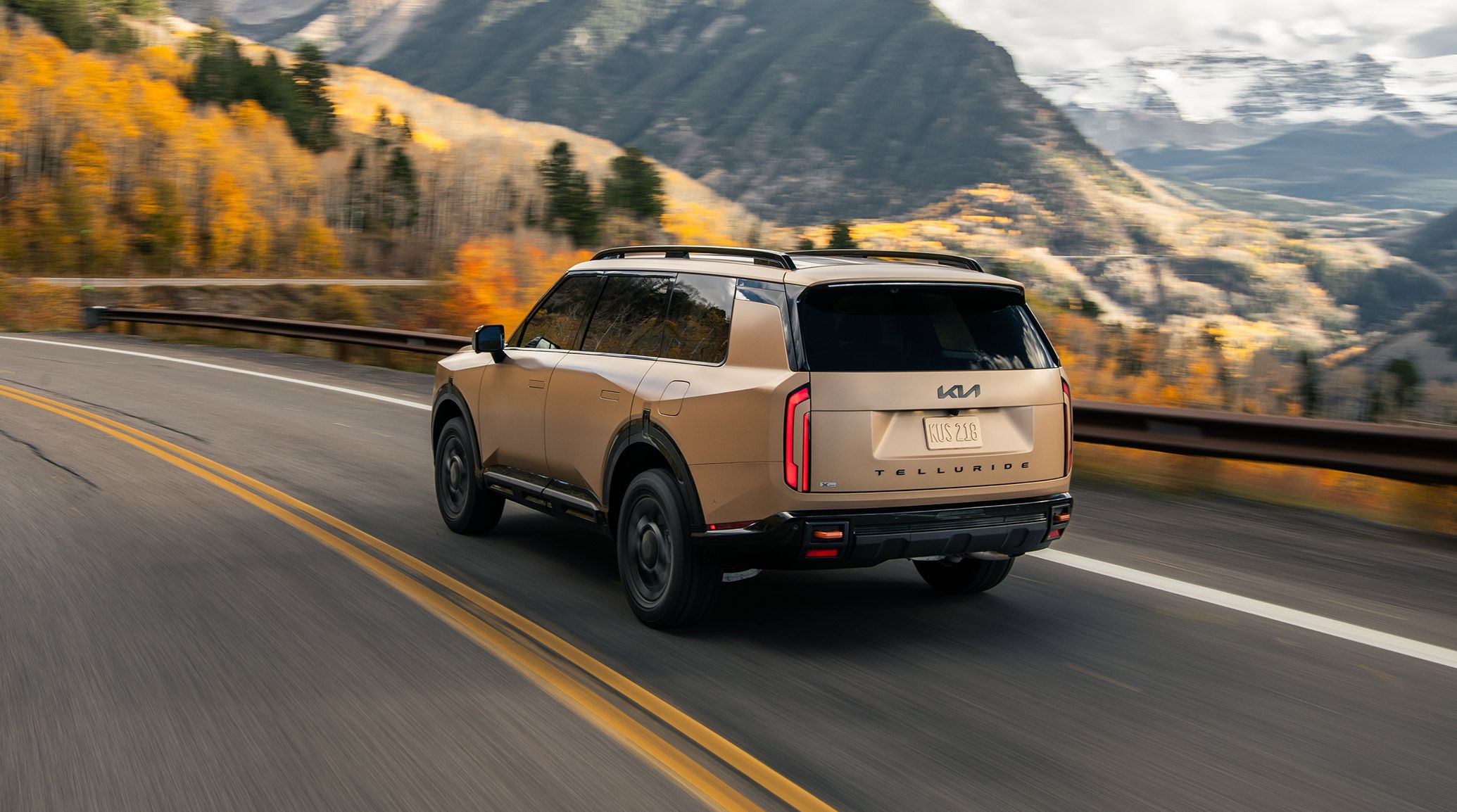 Rear view of 2027 Kia Telluride X-Pro SX Prestige in brown driving along a winding mountain road surrounded by vibrant fall plants and snow-capped peaks.