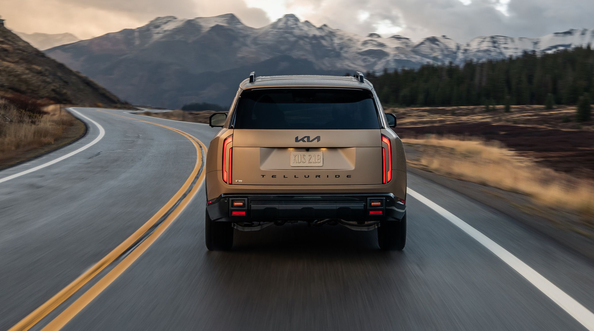 A rear-view image of the 2027 Kia Telluride SUV in brown driving along a winding mountain road, framed by dense forest and snow-capped mountains in the background.