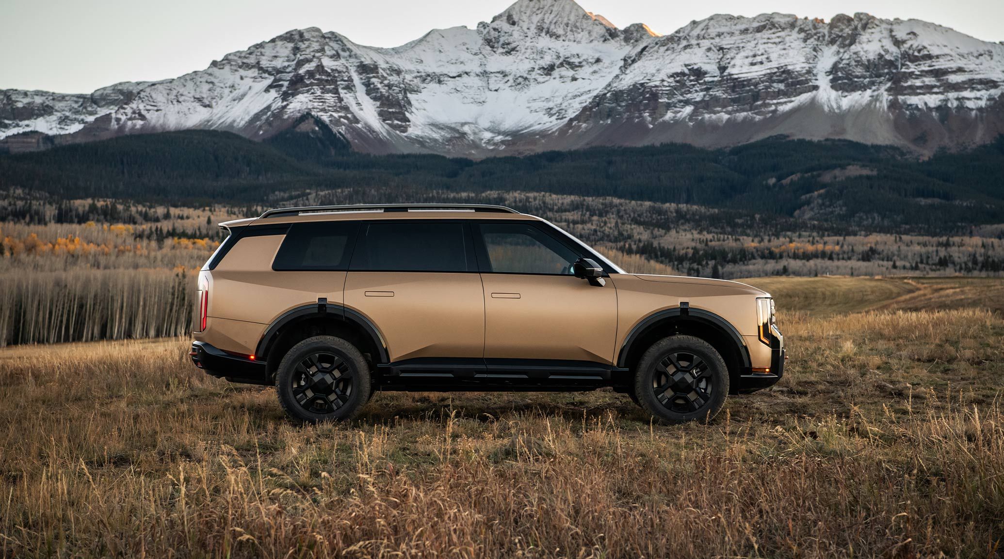 Side view of the 2027 Kia Telluride X-Pro in matte brown parked in a grassy field with snow-capped mountains in the background.