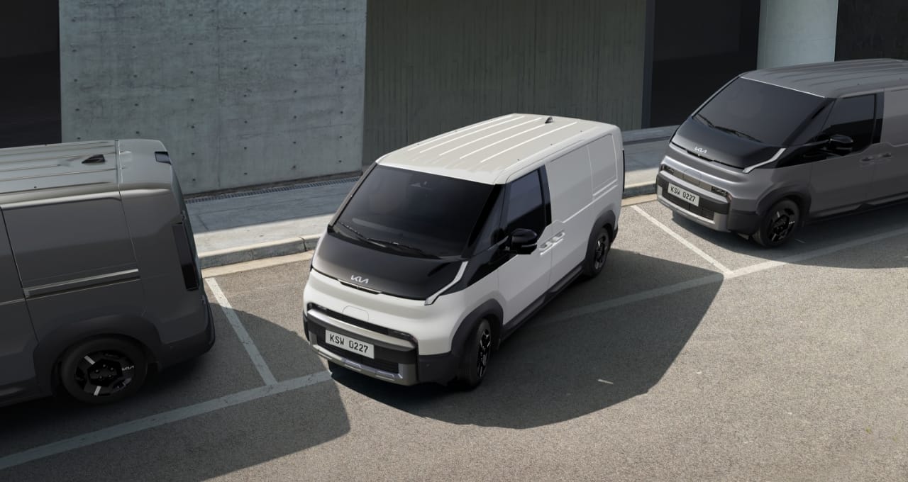 The Kia PV5 Cargo exiting parallel parking space, in a lot with two other vans