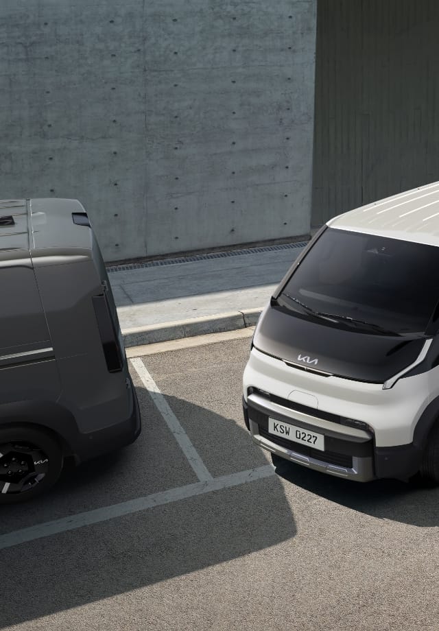 The Kia PV5 Cargo exiting parallel parking space, in a lot with two other vans
