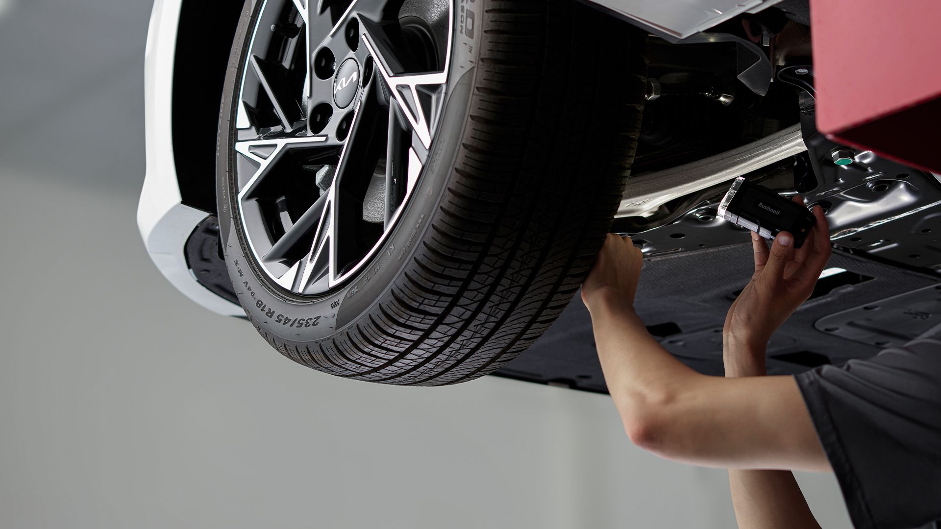 Technician inspecting the undercarriage of a lifted Kia K5 in a service bay