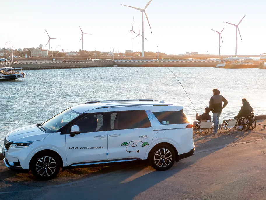 Kia mobility support vehicle parked at a waterfront near people using wheelchairs, with wind turbines in the background