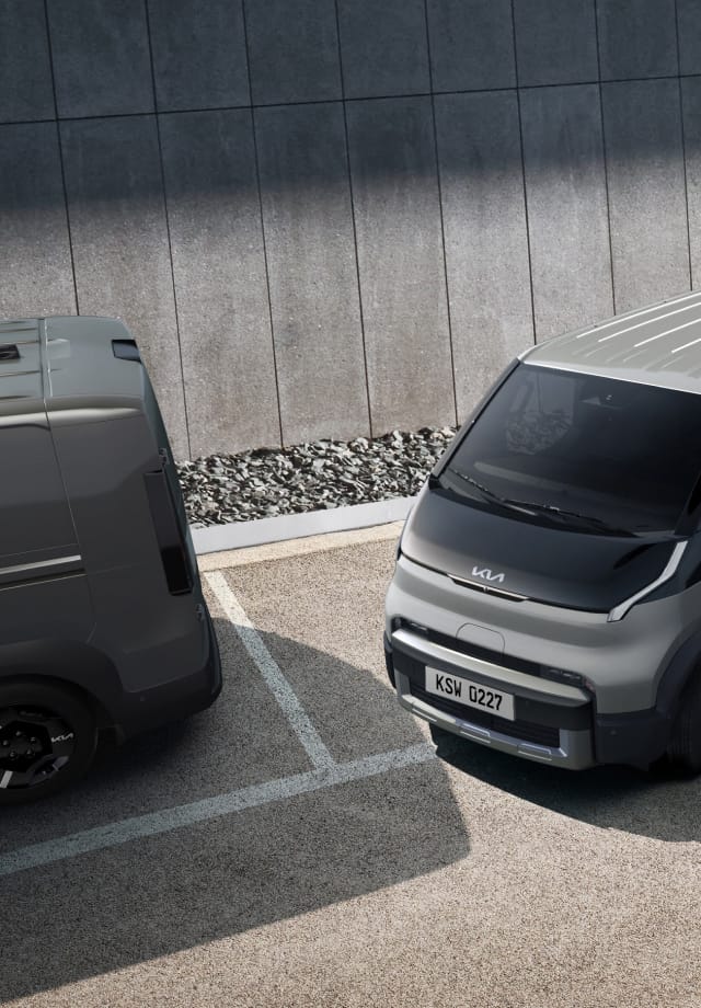 The Kia PV5 Cargo exiting parallel parking space, in a lot with two other vans