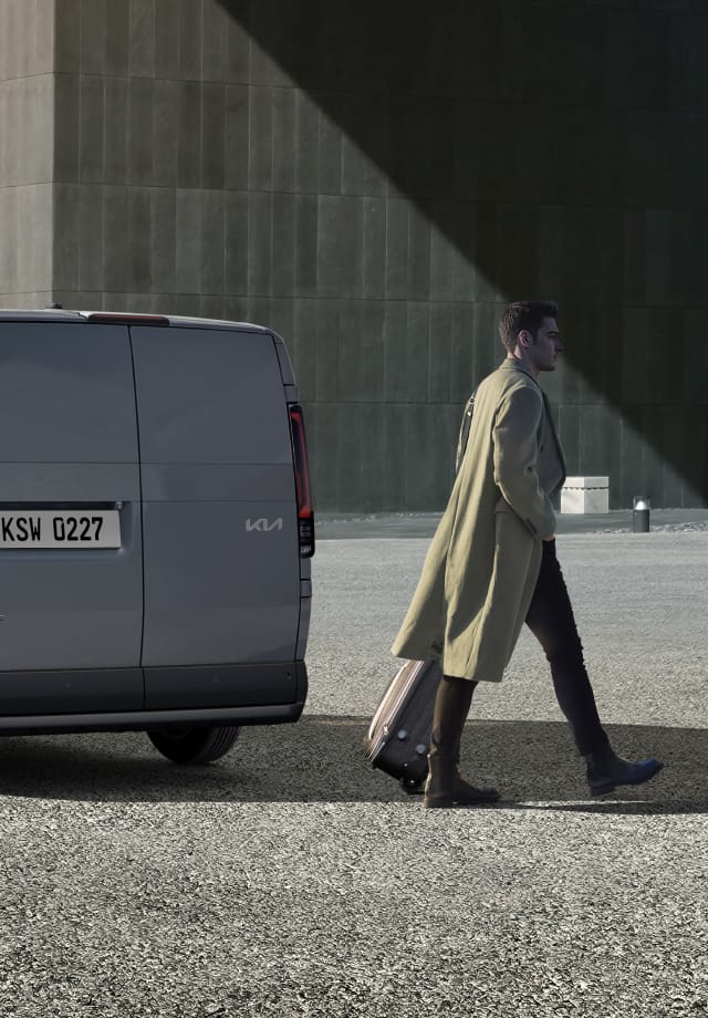 Rear three-quarter view of the Kia PV5 Cargo parked near a building with man walking by