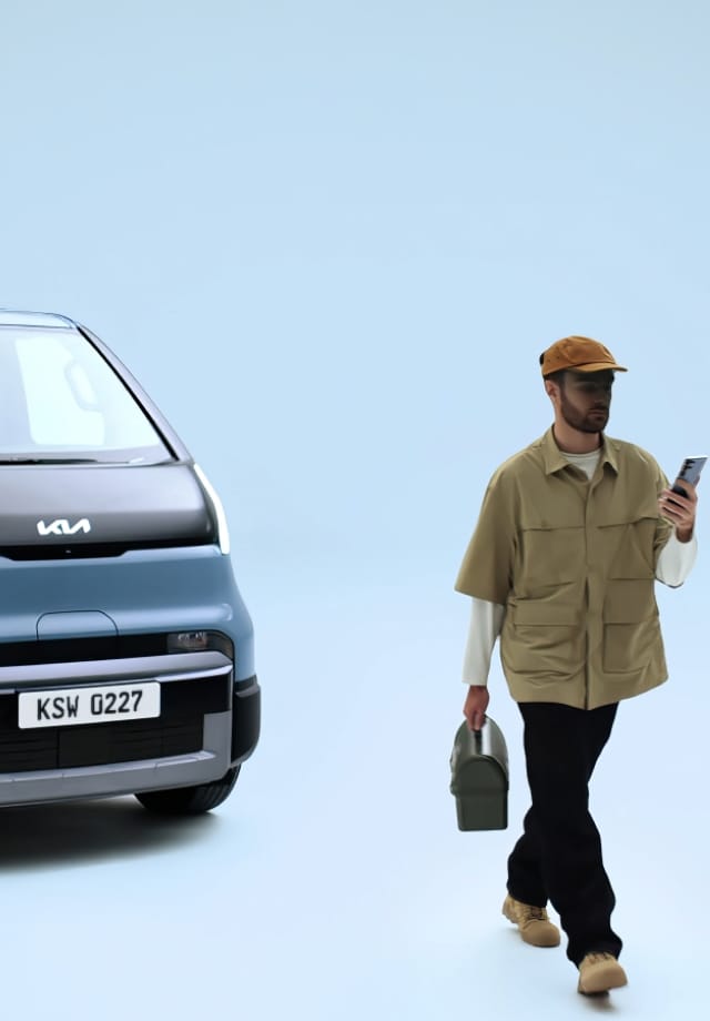 Hand holding a smartphone displaying a Kia PV5 digital key in front of the vehicle