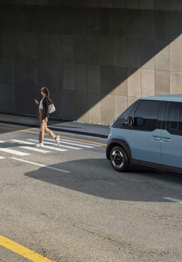 The Kia PV5 Passenger stopped at a crosswalk while a pedestrian walks by