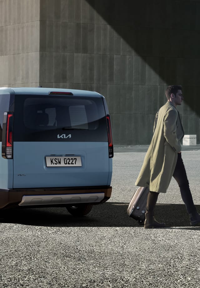 Rear three-quarter view of the Kia PV5 Passenger parked near a building with a person walking by
