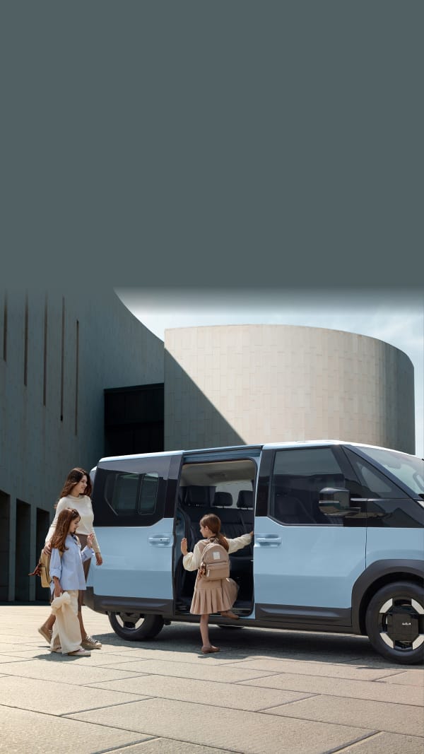 El Kia PV5 Pasajeros con dos puertas corredizas abiertas, mientras una madre y sus hijos se preparan para subir