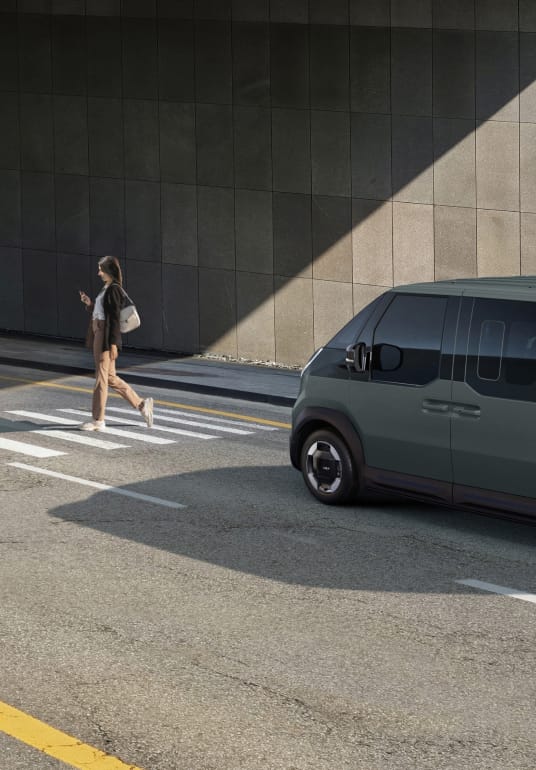 Kia PV5 WAV fährt auf einer Stadtstraße – der Frontkollisionswarner erkennt einen Fußgänger voraus.