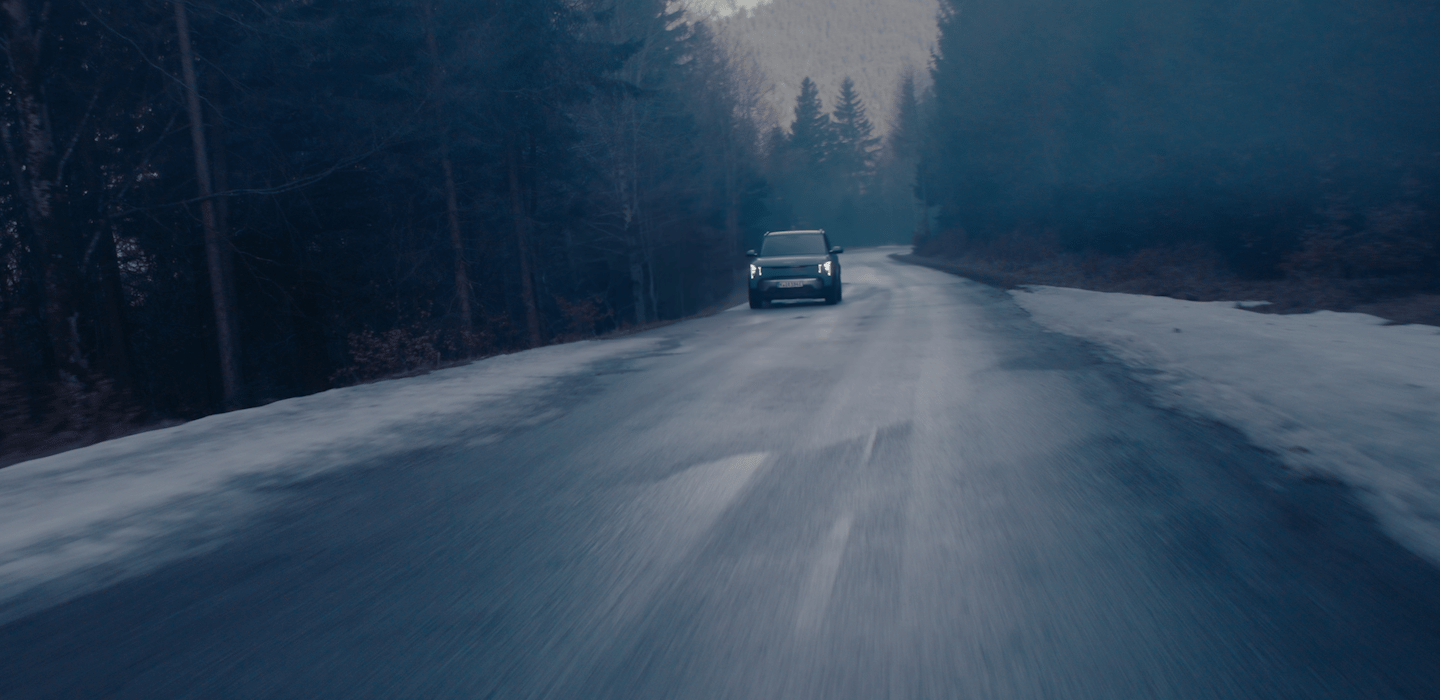 Dark, atmospheric view of a Kia vehicle driving toward the camera on a wet forest road at dusk, headlights on, with patches of snow along the roadside.