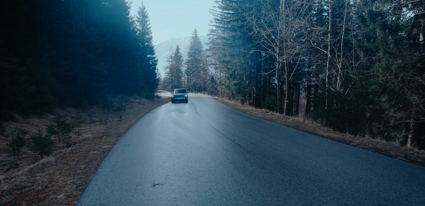 A Kia vehicle driving away from the camera down a forest road at dusk, framed by tall pine trees and distant mountains.