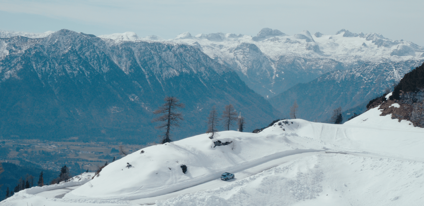 Aerial view of a Kia vehicle driving along a snow-covered mountain road, surrounded by dramatic snow-capped peaks.