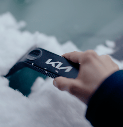 Close-up of a hand using a Kia-branded ice scraper to clear snow and ice from a car windshield.