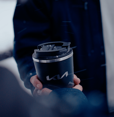 Close-up of hands holding a Kia-branded insulated travel mug outdoors in snowy weather.