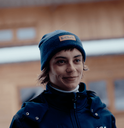 Person smiling outdoors wearing a Kia-branded beanie and navy jacket, with a wooden lodge in the background.
