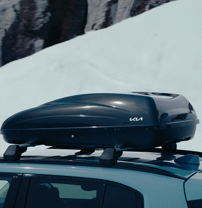 Kia vehicle with a rooftop cargo box mounted on roof rails, parked against a snowy mountain backdrop.