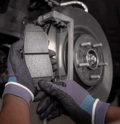 Close-up of a gloved technician's hands holding a brake pad next to an exposed brake disc and caliper during inspection.