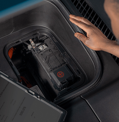 Close-up of a technician's hand near an open compartment revealing a vehicle's 12V battery and electrical components, with a diagnostic tablet showing battery percentage in the foreground