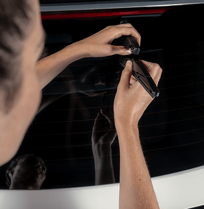 Close-up of a technician's hands installing a small component onto a vehicle's rear window, reflected against the glass.