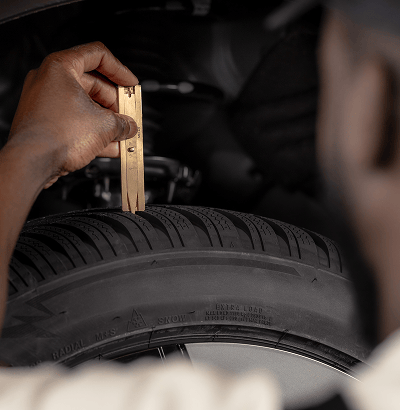 A technician in overalls inspecting a wiper blade component under an open car hood, with a turbocharged engine visible.