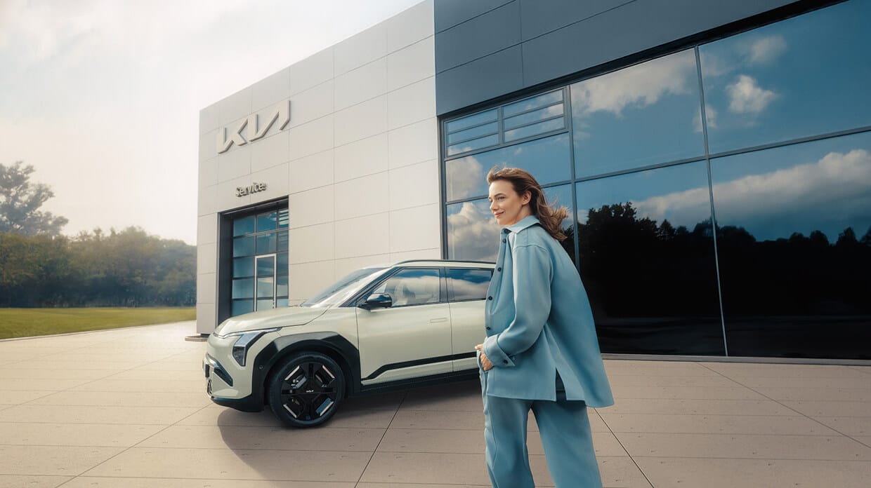 A woman is about to get into her freshly serviced Kia EV3 in front of a Kia dealership.