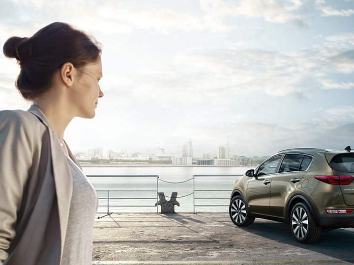 Una mujer mira al coche y al mar con una ciudad de fondo