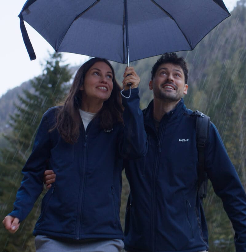 A Couple in Kia Softshell Jackets from the Kia Collection take cover under a Kia umbrella during a scenic mountain hike