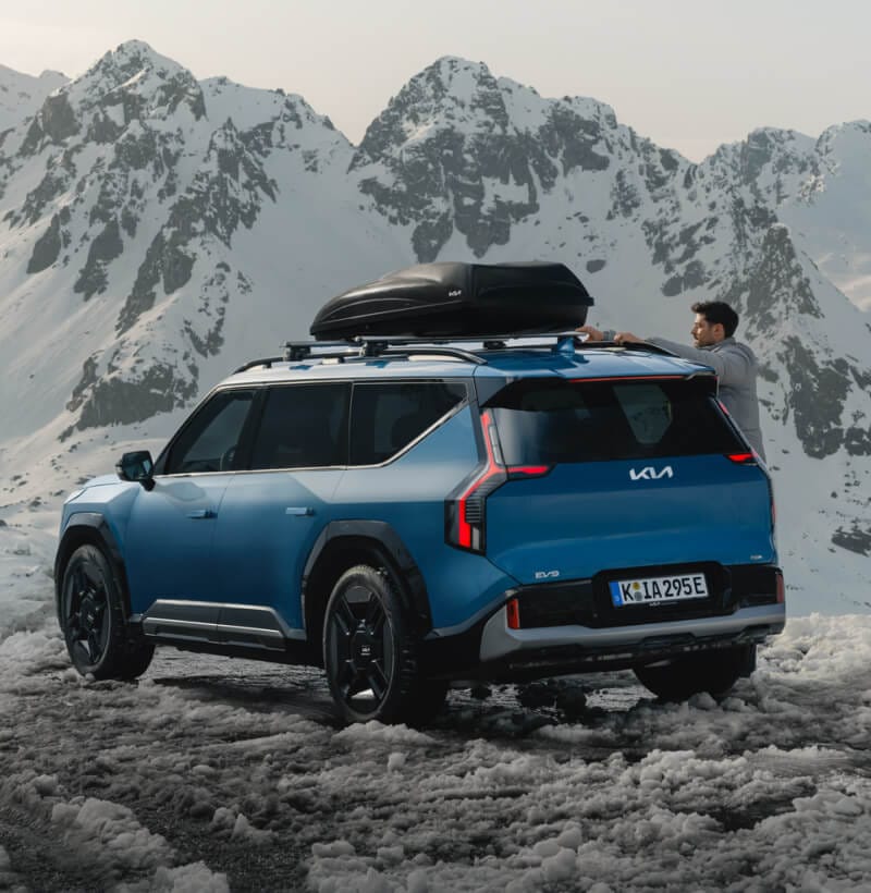 Kia EV9 with Kia Cross Bars and Kia Roof Box mounted in front of snowy mountains.