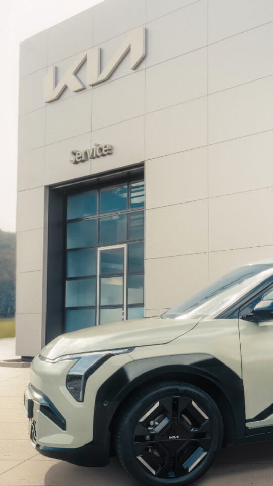 A woman is about to get into her freshly serviced Kia EV3 in front of a Kia dealership.