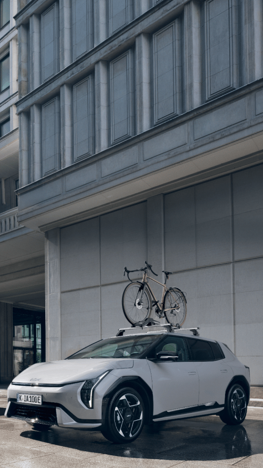 Two Kia EVs equipped with a Kia Roof Box and Bike Carrier Plus park in front of a building.