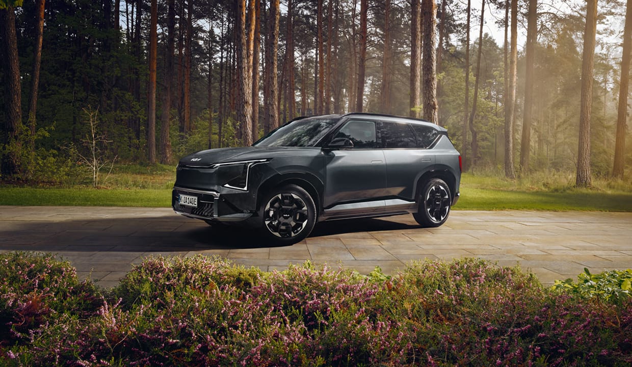 Kia EV5 stands outdoors surrounded by nature, with bright summer sunlight glinting off its glossy varnish.