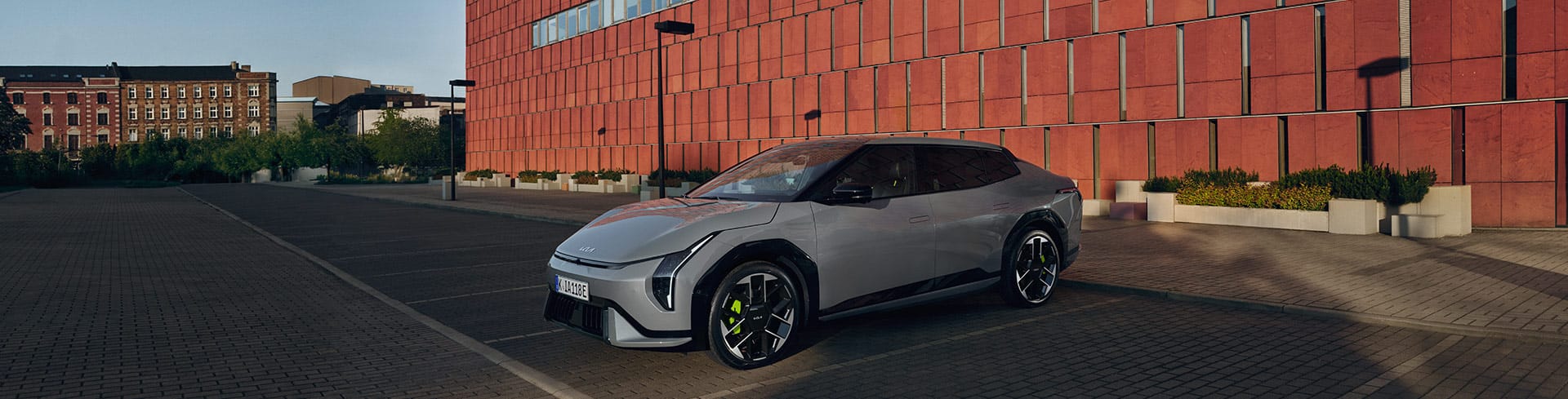 Vue extérieure de face de la Kia EV4 GT grise garée sur un parking pavé devant un grand bâtiment en briques rouges.