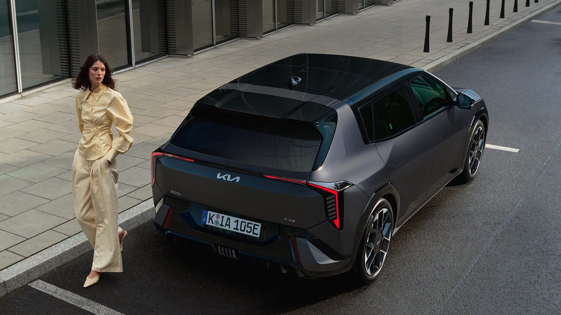 Une femme en tenue jaune pâle pose à côté d'une Kia EV4 GT gris foncé stationnée dans une rue moderne.