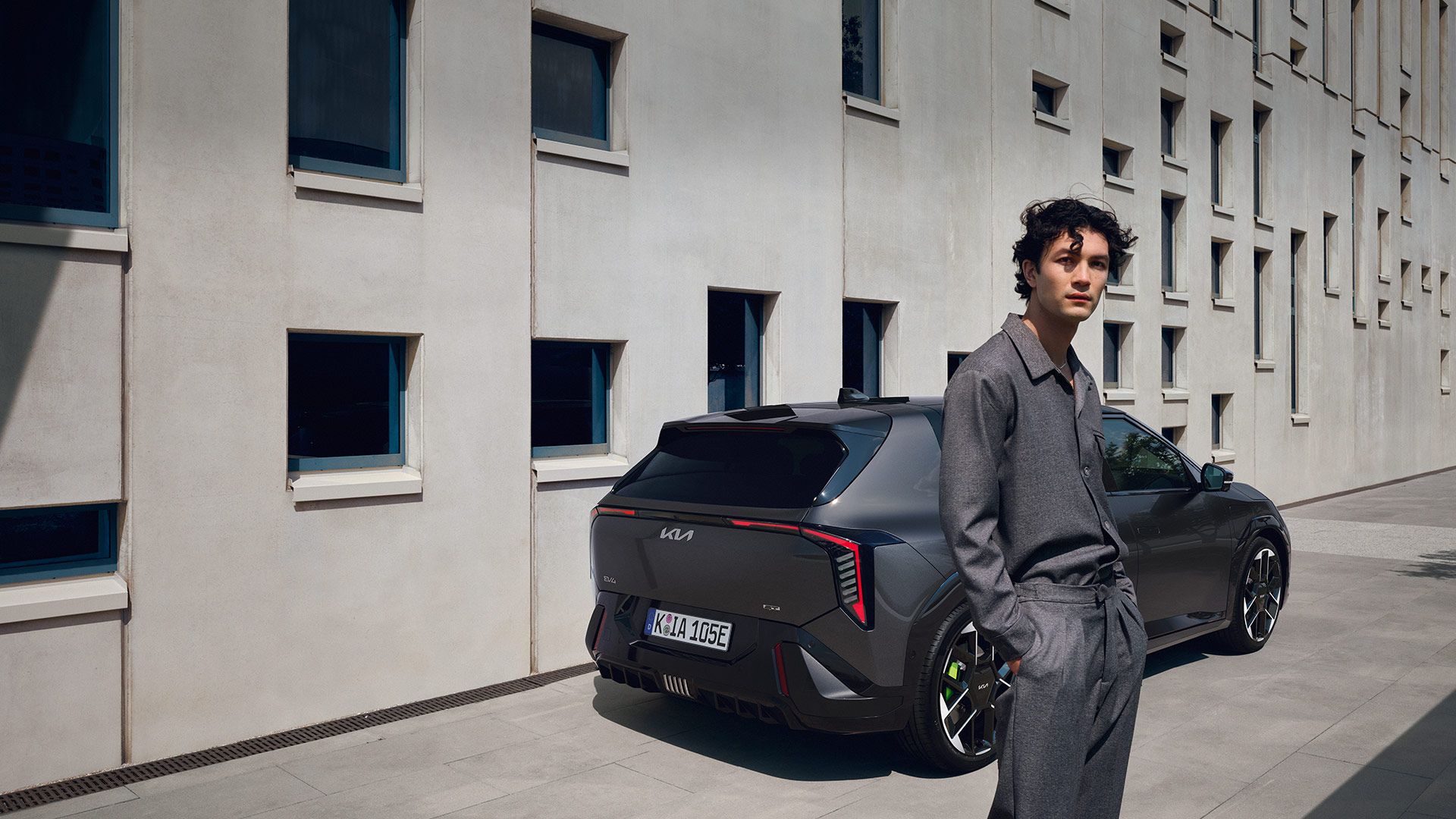 Homme debout au premier plan devant l'arrière d'une Kia EV4 GT gris foncé garée le long d'un bâtiment moderne