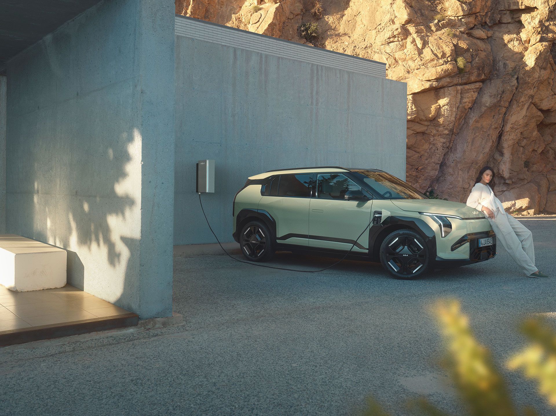 A person leans against a Kia EV3 charging at a station, set against a rocky mountainous background.
