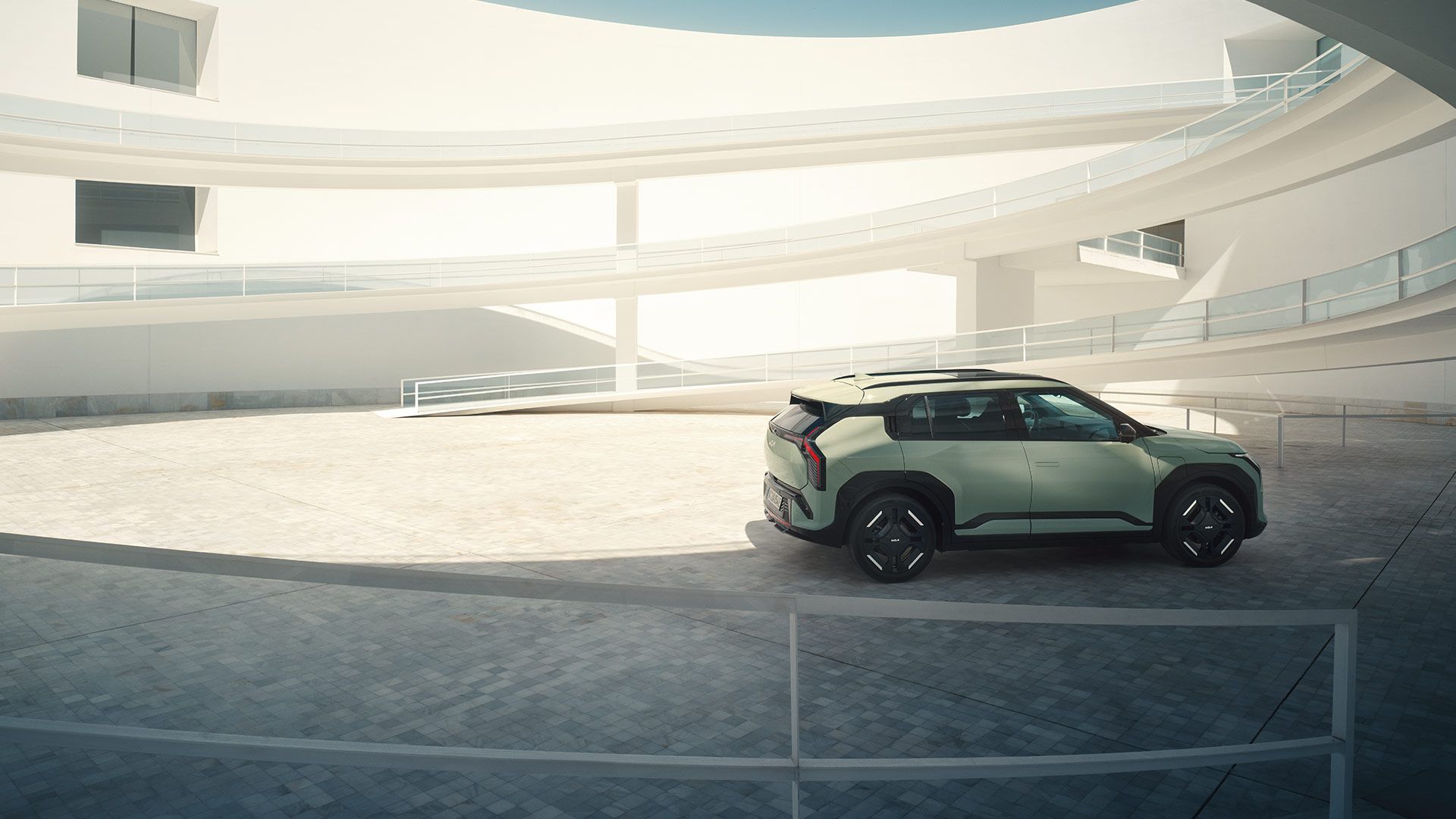 Rear-side view of a green Kia EV3 parked on the middle level of a sunny, modern white concrete car park