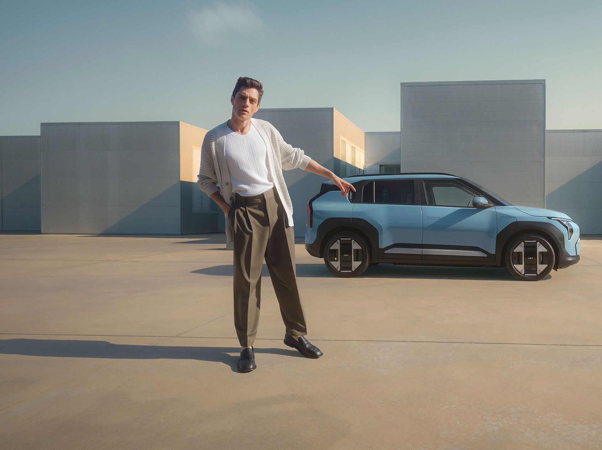 A person stands in front and pointd at a blue Kia EV3 parked in front of a contemporary panelled building