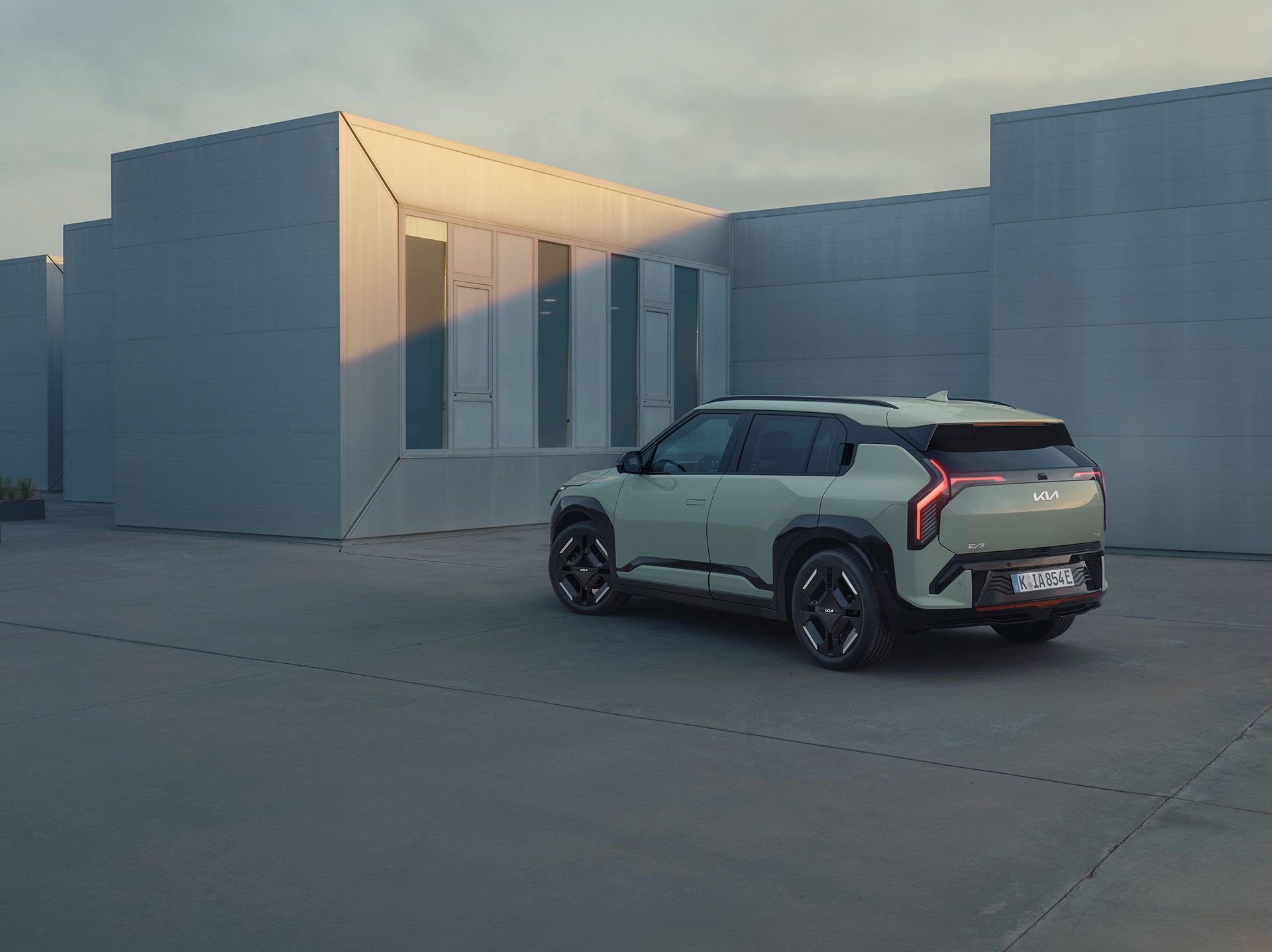 A Kia EV3 is parked on a concrete lot beside modern buildings under an overcast sky.